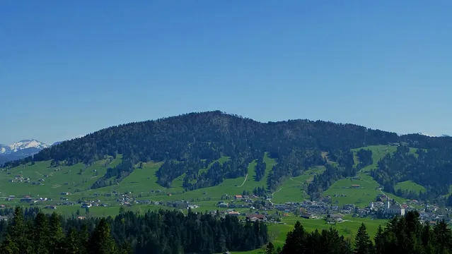 Landschaft mit grünen Hügeln, Dorf am Talboden und Waldgürtel, Berge im Hintergrund.