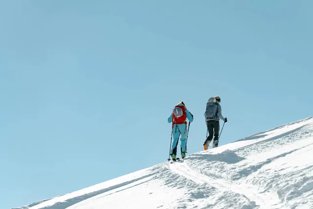 Zwei Skitourengeher steigen einen verschneiten Hang hinauf, blaue Jacken, Rucksäcke, Skier.