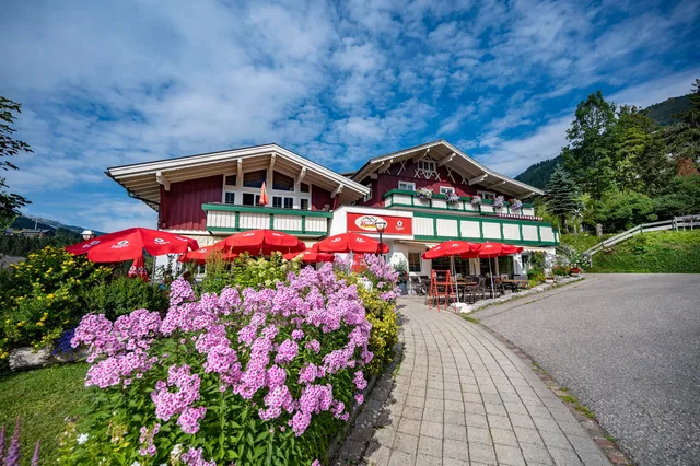 Alpenrestaurant mit roten Sonnenschirmen, Blumenbeet am Weg und Holzhütten im Alpenstil