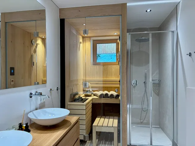 Modernes Badezimmer mit Holzdetails, Doppelwaschbecken, Glasdusche und Sauna im Hintergrund.