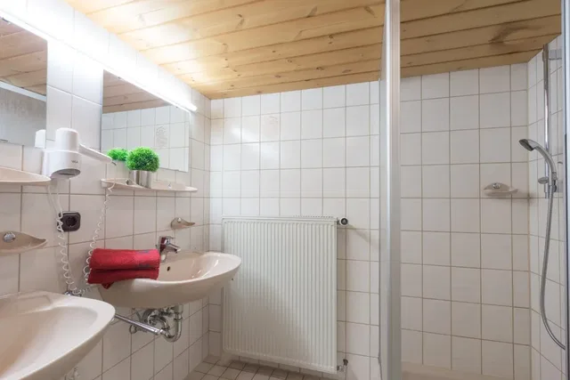 Kleines Badezimmer mit zwei Waschbecken, Spiegel, rotem Handtuch, Dusche und Heizkörper.
