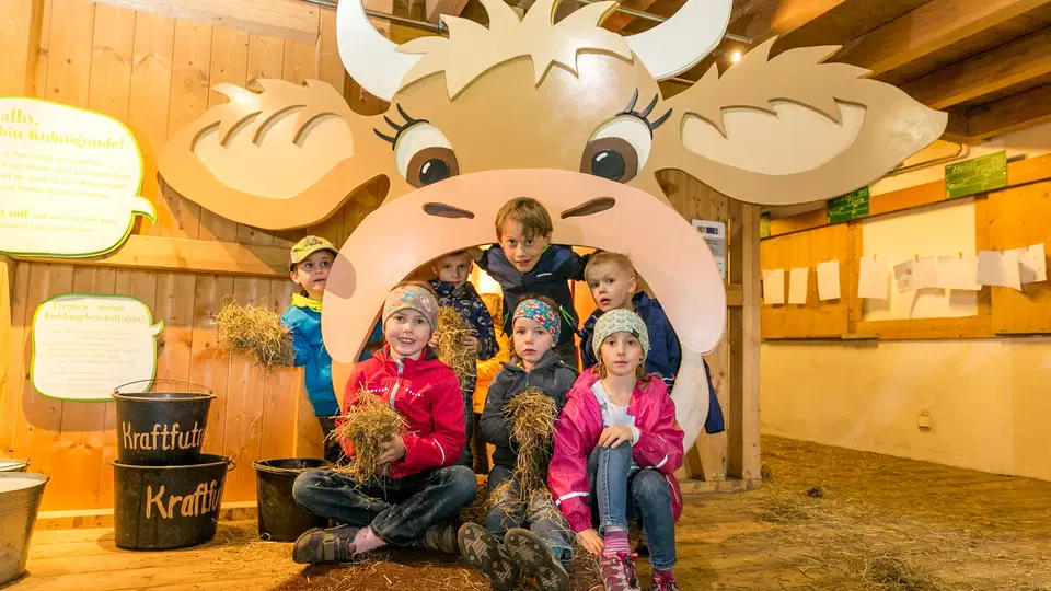 Gruppe Kinder sitzt unter einer großen Kuh-Attrappe im Stall