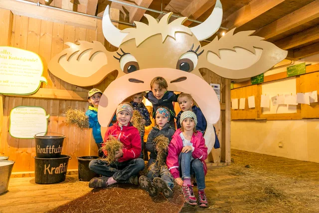 Gruppe Kinder sitzt unter einer großen Kuh-Attrappe im Stall