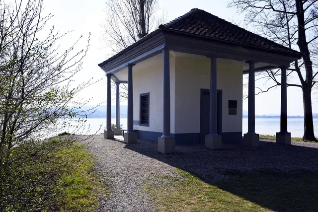Kleiner giebeldachiger Pavillon mit Säulen am Seeufer, umgeben von Bäumen.