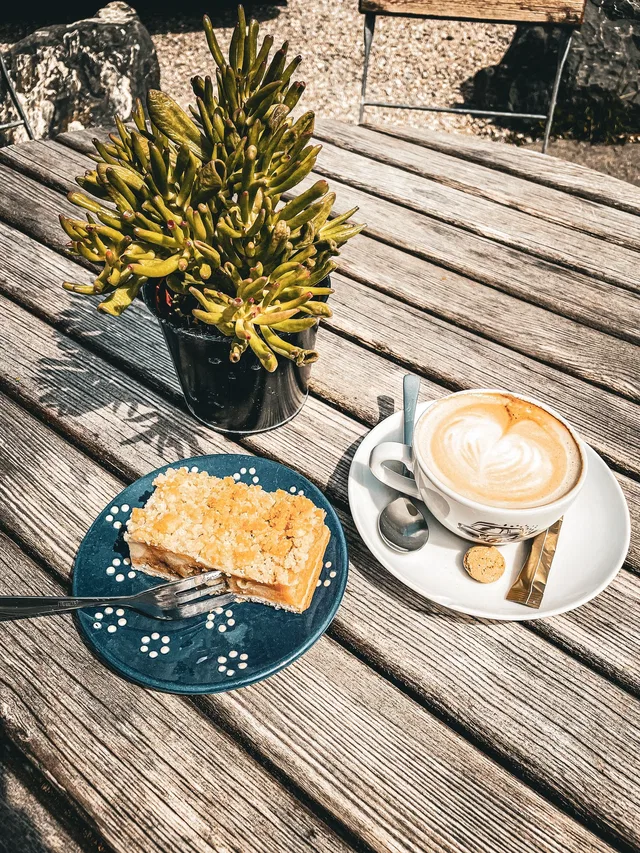 Kaffee mit Milchschaum, Kuchen auf blauem Teller und Topfpflanze auf Holztisch im Freien.