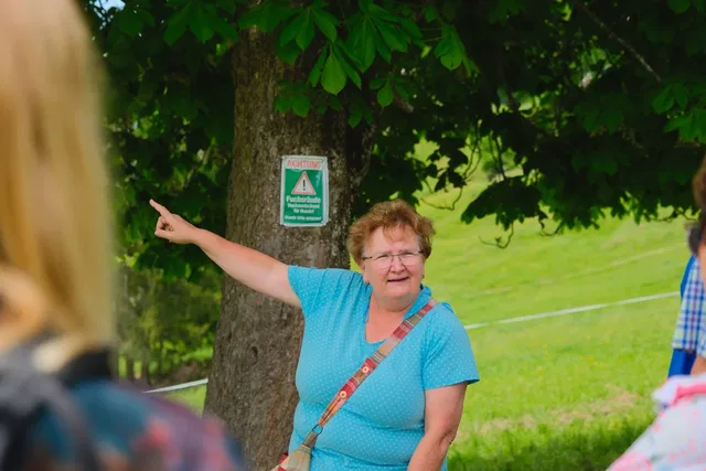 Frau in blauem Oberteil zeigt mit ausgestrecktem Arm neben einem Baum.