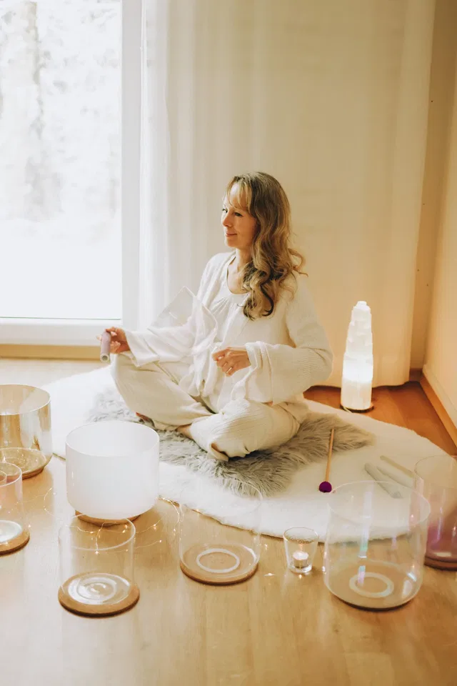 Frau sitzt im weißen Gewand auf Fell, Meditations-Setup mit Klangschalen im Raum.