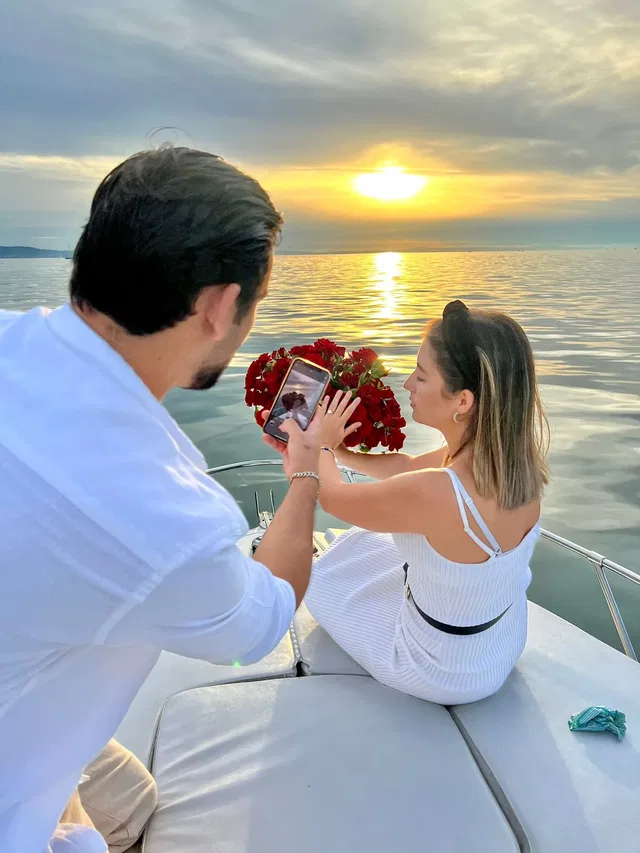 Paar auf Boot; Frau mit Rosenstrauß, Mann filmt sie bei Sonnenuntergang über dem Meer