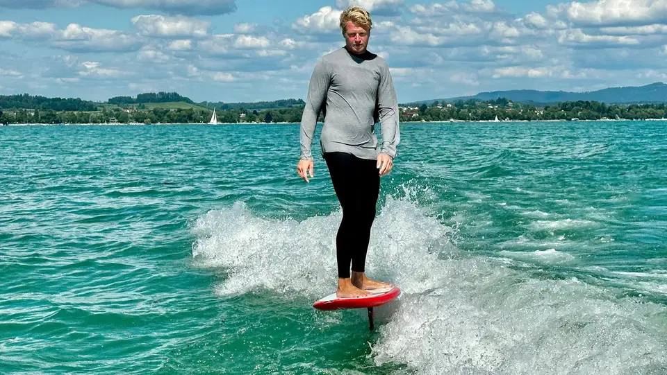 Mann steht auf Wakefoil-Board auf dem Wasser, sprayt Wasserspritzer um ihn