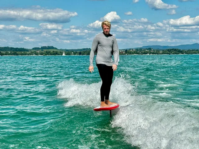 Mann steht auf Wakefoil-Board auf dem Wasser, sprayt Wasserspritzer um ihn
