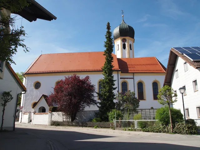 Historische Kirche mit rotem Dach und grünem Zwiebelturm, von Bäumen und Häusern umgeben.
