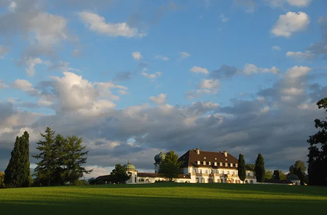 Historische Schlossanlage auf grünem Gelände mit Bäumen und Wolkenhimmel.