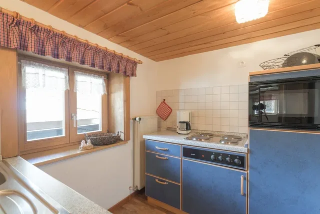 Küche mit blauen Fronten, Holzdecke, Fenster mit kariertem Vorhang, Herd, Ofen und Spüle.