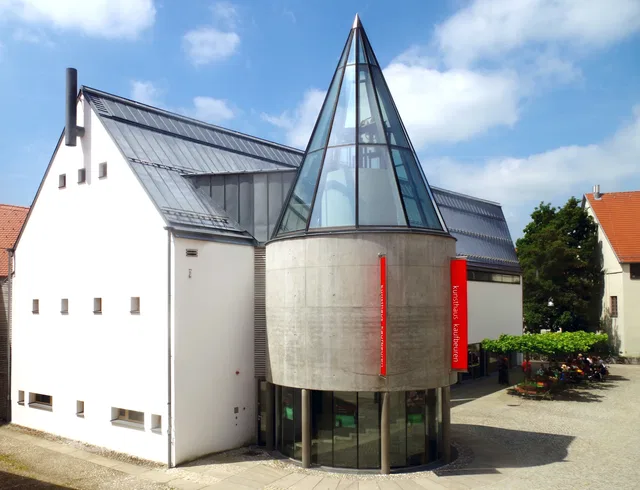 Außenansicht des Kunsthauses Kaufbeuren mit gläsernem Turm und weißem Gebäudekomplex.