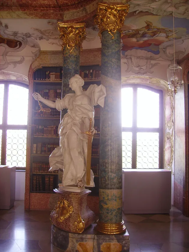 Weiße Skulptur einer bewaffneten Frau zwischen vergoldeten Säulen und Bücherregalen