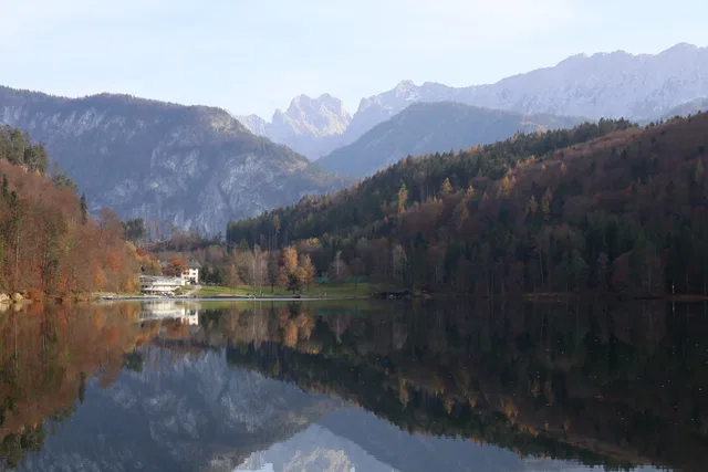 Bergsee mit Herbstbäumen, Ufergebäude und spiegelnder Berglandschaft im Hintergrund.