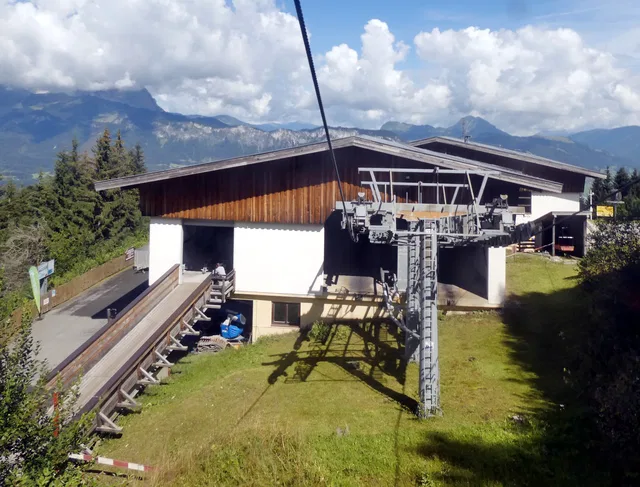 Skiliftstation auf einer grünen Wiese mit Seilbahn-Turm vor Bergen.
