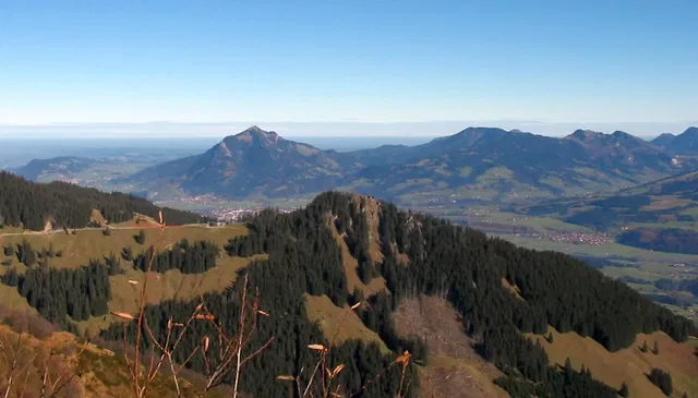Berglandschaft mit bewaldeten Hängen, grünem Talboden und Dörfern im Hintergrund unter blauem Himmel