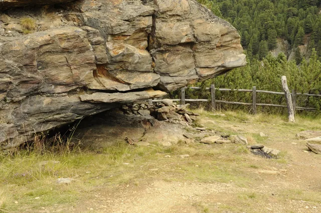 Große Felswand über einem Wanderweg, Holzzaun im Hintergrund.
