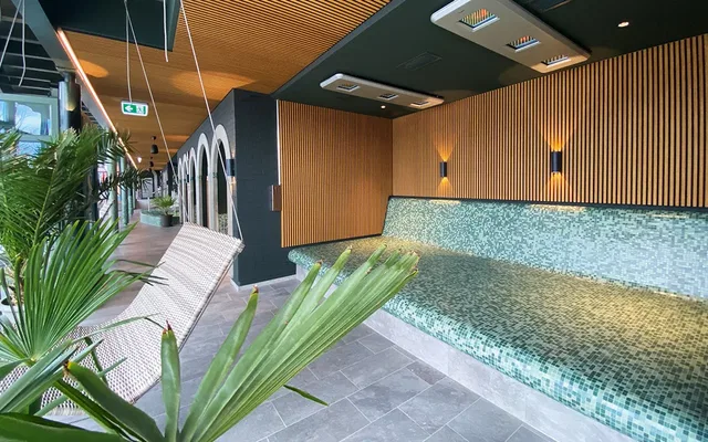 Thermalbereich mit mosaikverkleideter Sitzbank, Holzlatwand und Grünpflanzen im Vordergrund.
