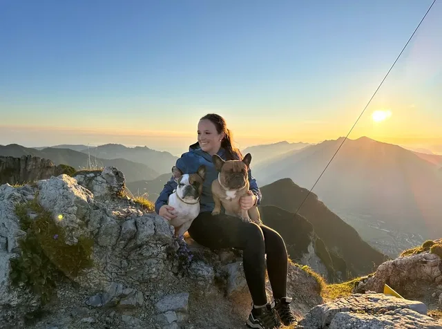 Frau sitzt auf Felsen mit zwei Hunden vor Bergpanorama bei Sonnenuntergang.