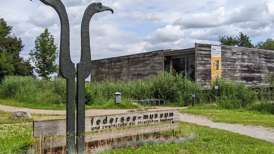 Zwei lange Vogelskulpturen vor einem holzverkleideten Museumsgebäude auf grünem Gelände.