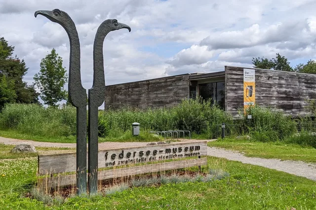 Zwei lange Vogelskulpturen vor einem holzverkleideten Museumsgebäude auf grünem Gelände.