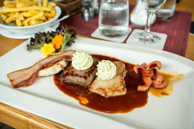 Fleischplatte in Bratensauce mit Kräuterbutter-Tupfen, Speckscheiben und Garnitur auf weißem Teller.