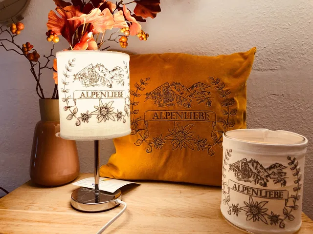 Gelbes Alpenliebe-Kissen neben einer Lampe und weißen Dekoobjekten mit Bergmotiven.