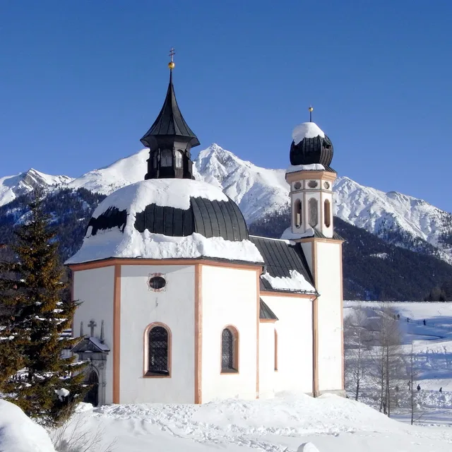 Weiße Kirche mit schwarzen Kuppeln und verschneitem Dach vor schneebedeckten Bergen.