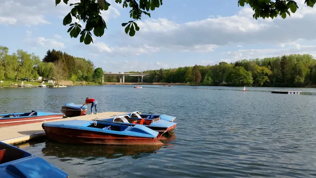 Boote am Ufer eines ruhigen Sees mit Brücke im Hintergrund und Bäumen am Ufer.