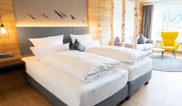 Hotelzimmer mit zwei zusammengestellten Betten, Holzdekoration, Bergbild und gelbem Sessel im Sitzcö