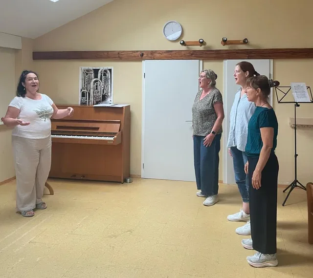 Frau spricht zu vier stehenden Frauen in einem Probenraum mit Klavier.
