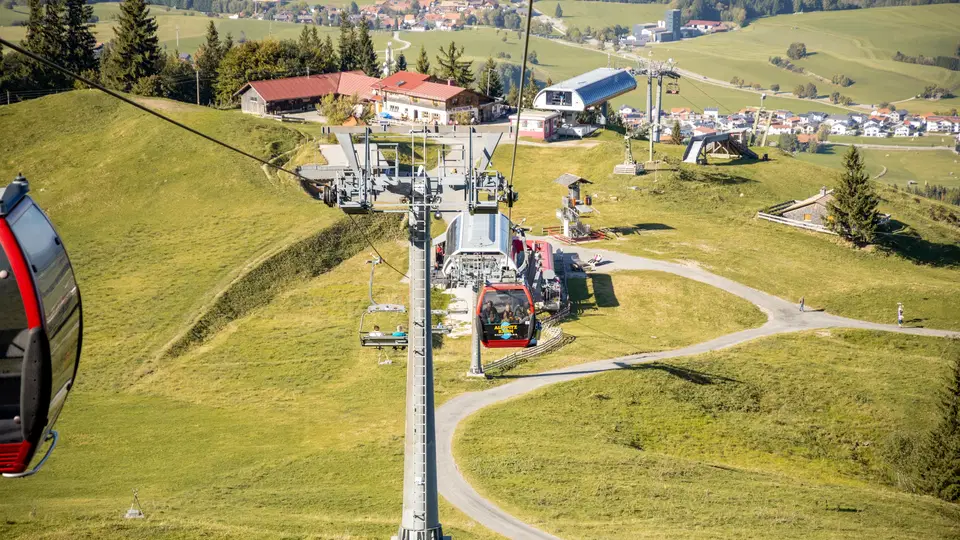 Seilbahngondeln fahren am Berghang vorbei über grüne Wiesen und Gebäude.