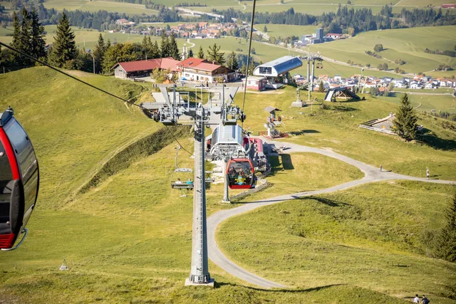 Seilbahngondeln fahren am Berghang vorbei über grüne Wiesen und Gebäude.