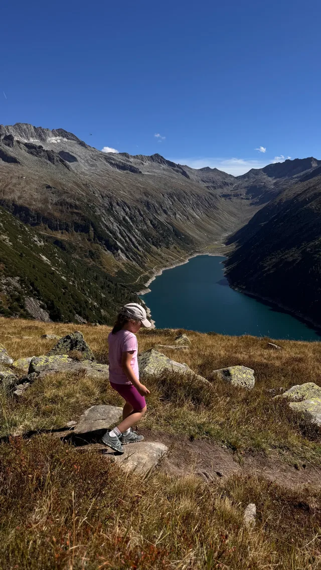 Ein Mädchen in Rosa Kleidung und Hut wandert auf einem felsigen Hang mit Blick auf einen Bergsee.