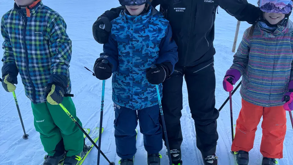 Vier Kinder mit Skiausrüstung und eine Begleitperson stehen auf der Piste mit Skiern, Berglandschaft