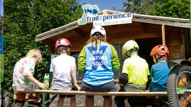 Kinder mit Helmen sitzen an einer Bank vor der TrailXperience-Hütte