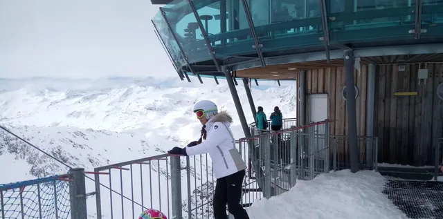 Person in weiße Winterjacke mit Helm am Geländer einer Bergbahn im Schnee.