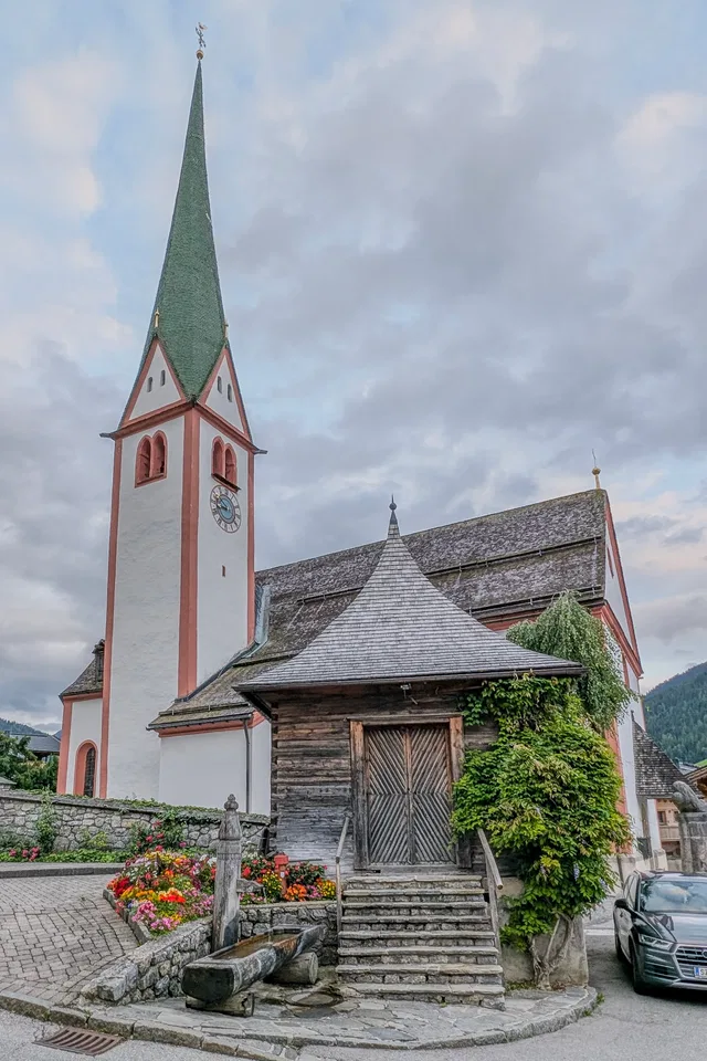 Historische Kirchturmspitze mit grünem Dach, daneben Holzbau und Blumengarten.