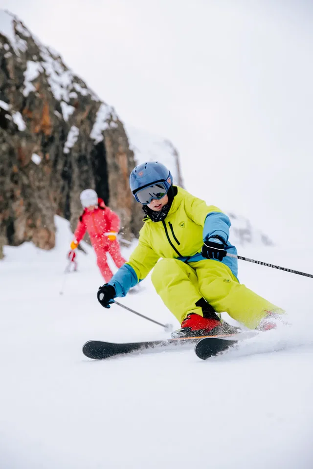 Kleiner Skifahrer in limegrüner Jacke und blauer Ausrüstung beim Kurvenfahren im Schnee.