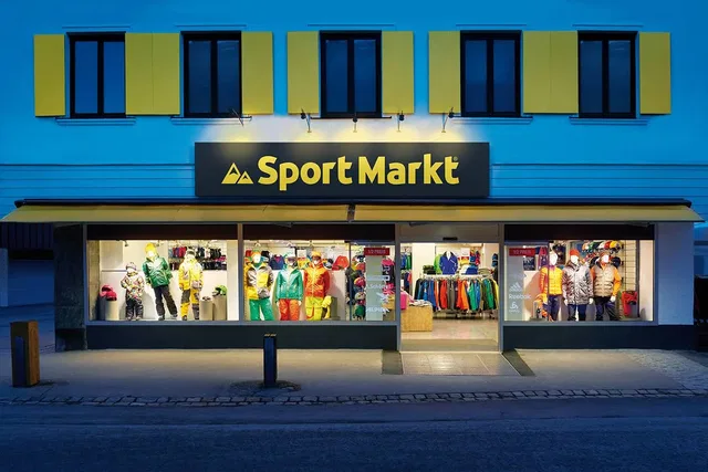 Außenansicht eines Sportmarkts mit gelben Fensterläden und beleuchtetem Schild 'Sport Markt' bei Däm
