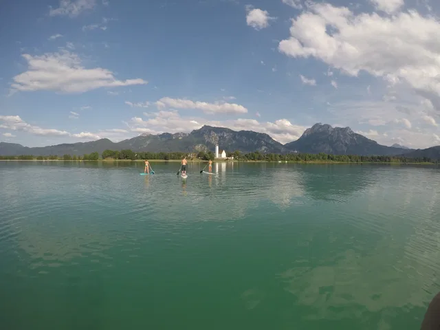 Paddleboarder auf ruhigem See vor Bergkulisse und blauem Himmel.