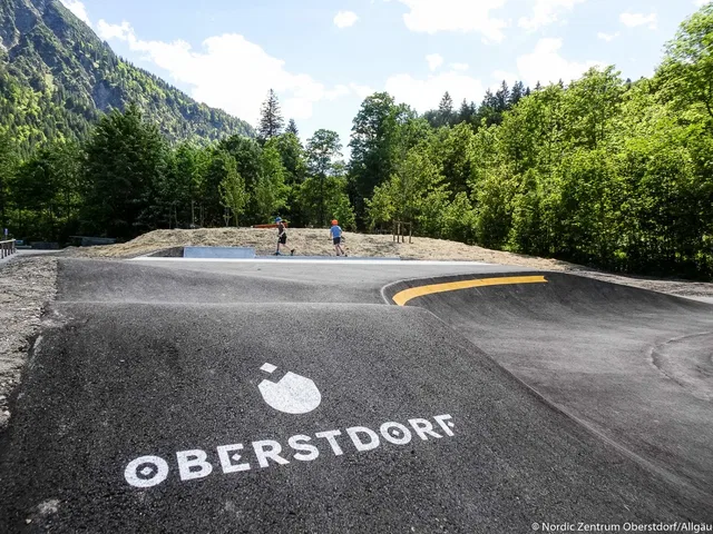 Pumptrack in Oberstdorf mit zwei Personen am Rand und bewaldeter Berglandschaft.