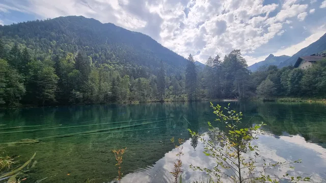 Klarer Bergsee umgeben von Wald, Berge im Hintergrund, Wolken am Himmel.