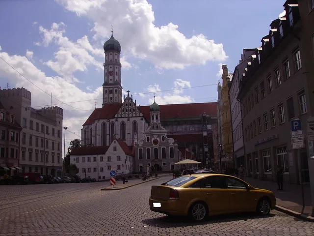 Historische Kirche mit hohem Turm an einem gepflasterten Stadtplatz, davor ein gelbes Taxi