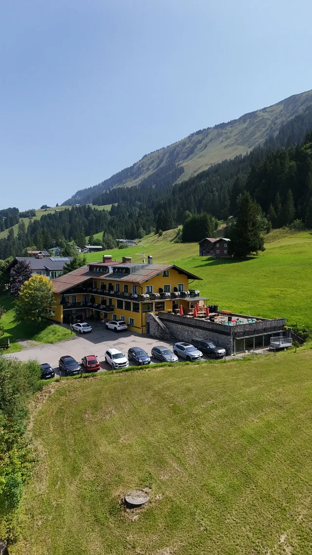 Gelbes Hotelgebäude am Hang mit Parkplätzen davor und umliegenden Wiesen