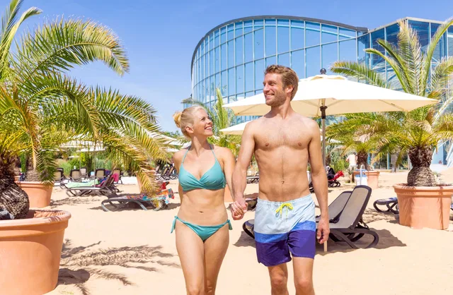 Paar in Badekleidung geht am Strand vorbei, Palmen und moderne Glasfassade im Hintergrund.
