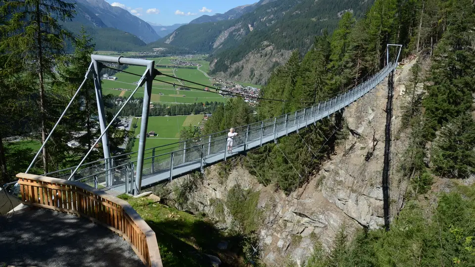 Symbolbild: Hängebrücke über steilem Felsabhang mit Blick auf Tal und Dorf in der Berglandschaft Keine Originalaufnahme dieses Eintrags.