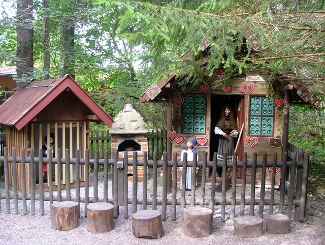 Kleines Waldspielhaus mit Zaun, Herzdeko und Personen davor.